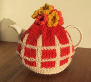 Red tea cosy with white picket fence detail and flowers on top